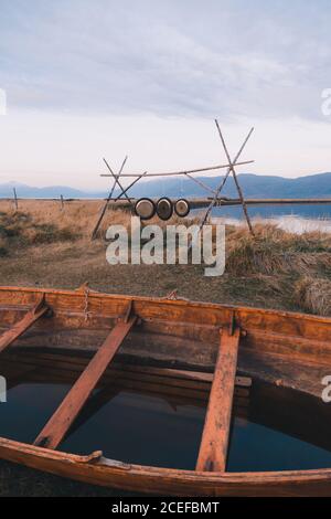 View of wooden old boat filled with water on cold shoreline with wooden construction and metal discs Stock Photo