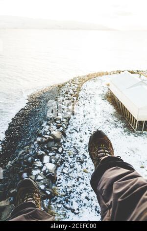 Crop legs of traveler in boots above rocky coast in snow with tent and water in bright light Stock Photo