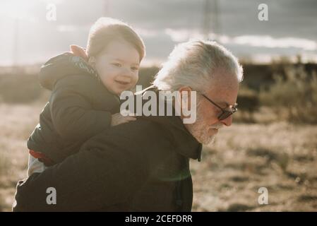 Side view of serious elderly man in sunglasses giving adorable infant piggyback ride in sunlight. Stock Photo