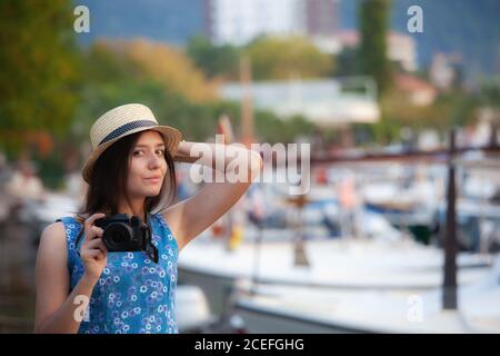 Lovely young Woman in stylish hat holding photo camera while standing on blurred background of harbor Stock Photo