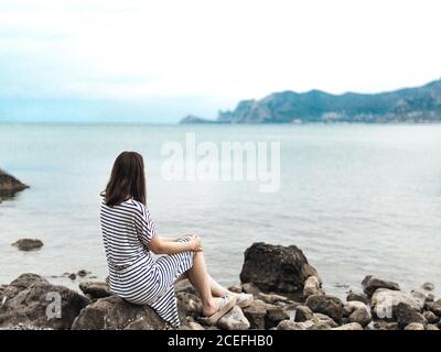 Back view of Woman in striped dress sitting alone on rocks of seashore looking away in dreams Stock Photo