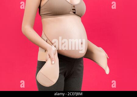 Close up of pregnant woman putting on a bandage at pink background with copy space. Orthopedic abdominal support belt concept. Stock Photo