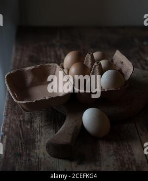 Carton with fresh chicken eggs lying on cutting board on wooden table in dark room Stock Photo