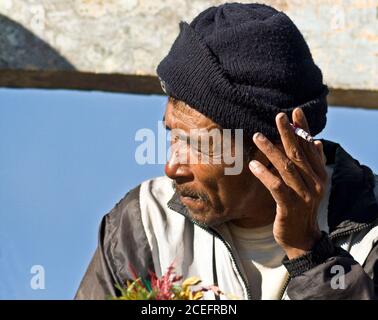 Indonesia - June 9, 2009: Side view of old poor male sitting, smoking and holding plants near woman behind Stock Photo