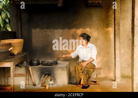 Indonesia - June 9, 2009: Old female in white shirt and colored skirt sitting on seat near burning furnace and mixing in frying pan by skimmer Stock Photo