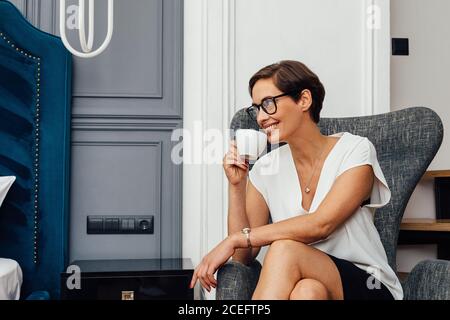 Smiling woman in casuals sitting on armchair and holding a cup. Side view of female entrepreneur drinking coffee in an apartment. Stock Photo