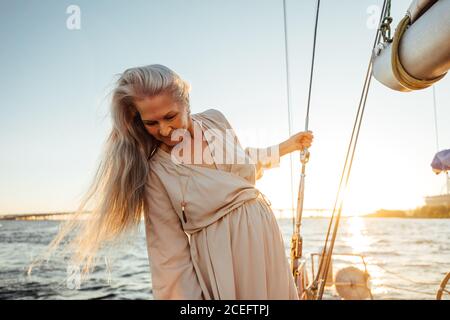 Beautiful mature woman with long gray hair standing on a sailboat and looking down at sunset Stock Photo