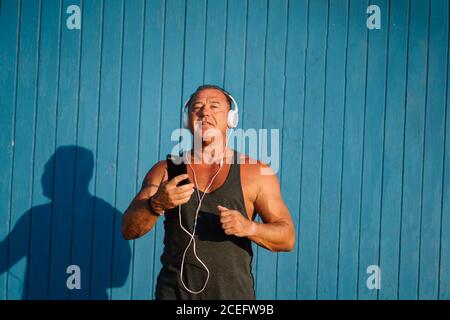 Strong older man poses with headphones on blue background. Stock Photo