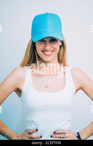 Young beautiful sportsWoman in white top and blue cap standing in studio with hands on hips and looking at camera on white background Stock Photo