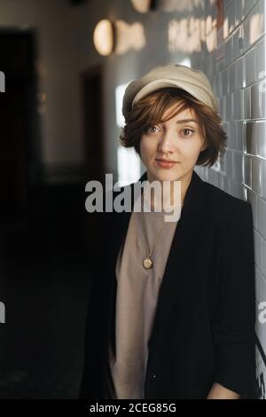 Young Woman with short hairstyle wearing simple black jacket and beige cap looking at camera and leaning on wall Stock Photo