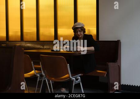 Young pretty Woman in cap sitting alone at table in modern restaurant and reading through menu Stock Photo