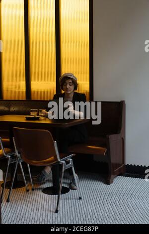 Young pretty Woman in cap sitting alone at table in modern restaurant and reading through menu Stock Photo