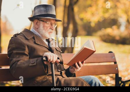 Profile side photo of attentive concentrated old man rest relax autumn town trees park read encyclopedia book sit bench hold walk stick wear cap Stock Photo