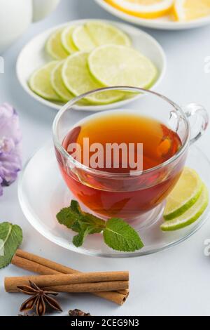 Beautiful set of hot black tea in glass cup on saucer placed on white background with brown sugar, cinnamon, leaves of mint, flowers and slices of citrus fruits on saucers with teapot behind Stock Photo