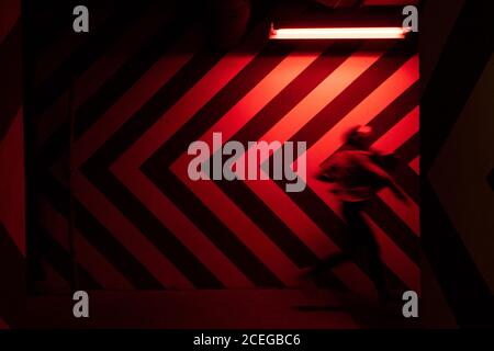 Side view of motion blurred figure of male walking down in tunnel in direction opposite to large red and black arrows on wall lit by red lamps Stock Photo