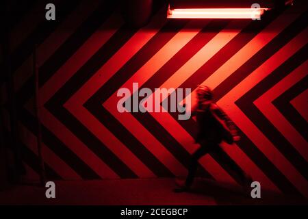 Side view of motion blurred figure of male walking down in tunnel in direction opposite to large red and black arrows on wall lit by red lamps Stock Photo