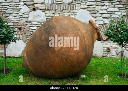 Traditional Georgian terracotta kvevri for making wine lying on grass between small tree saplings on background of white stone and brick wall Stock Photo