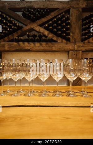Row of elegant shiny glasses standing on wooden table in wine cellar with wine bottles on shelves on background Stock Photo