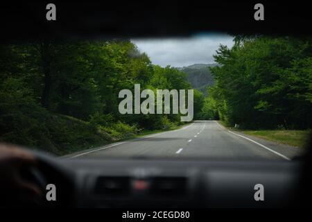 View through car windscreen of scenic highway leading through forest to distant mountains on cloudy day Stock Photo