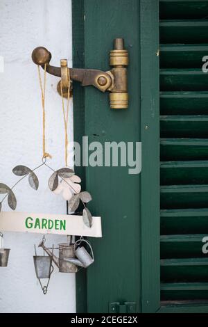 Close-up view of old rustic green wooden door with worn bronze latch and beautiful metal garden sign with watering can and bucket figurines hanging from it at white plastered wall Stock Photo