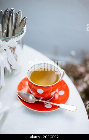 Red ceramic polka-dotted mug on saucer with spoon?and light drink in it standing on white round table with elegant jars with lace ribbons outdoors from above Stock Photo