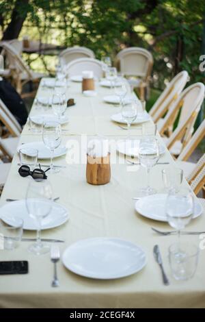 Beautiful table with light cloth and rattan chairs around set with empty wine glasses and plates outdoors on pavement with trees and grass around Stock Photo