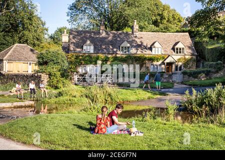 Visitors enjoying the evening sunlight beside the ford across the River Eye in the Cotswold village of Upper Slaughter, Gloucestershire UK