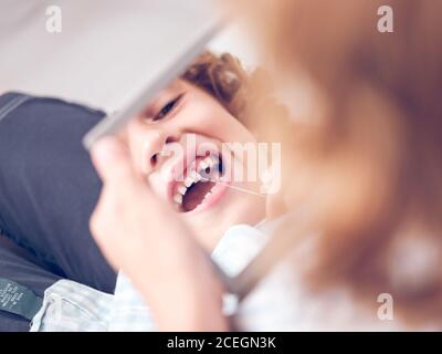 Crop little boy holding small mirror and looking in reflection while pulling out tooth with thread Stock Photo