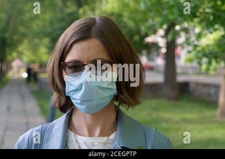 Young woman in medical mask standing in city park looking to camera green woods on background Concept of health and safety life COVID-19 coronavirus v