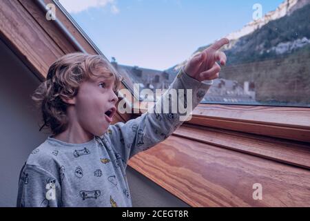 Cute little boy in pajamas pointing at wonderful mountain while looking out window of countryside house Stock Photo