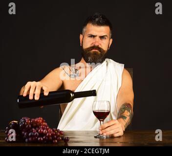 Man with beard holds glass of wine on brown background. Viticulture and crops concept. Sommelier pours expensive alcohol to taste. God Bacchus with strict face wearing white cloth sits by grapes Stock Photo