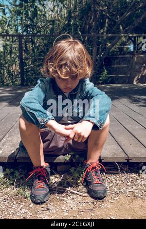 Cute little boy with curly blond hair sitting on small wooden bridge looking down in countryside Stock Photo