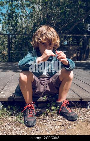 Cute little boy with curly blond hair sitting on small wooden bridge looking down in countryside Stock Photo