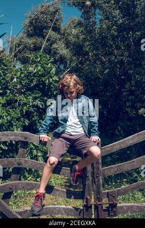 Cute little boy with curly blond hair sitting on small wooden bridge looking down in countryside Stock Photo