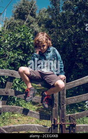 Cute little boy with curly blond hair sitting on small wooden bridge looking down in countryside Stock Photo