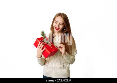 Surprised young woman holds in a white sweater holding a Christmas red gift with a fir branch and a festive bow. The happy model is happy to receive a gift and opens her mouth in surprise. Stock Photo