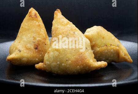 Three golden brown samosas on a black plate. Popular Indian snack. Stock Photo
