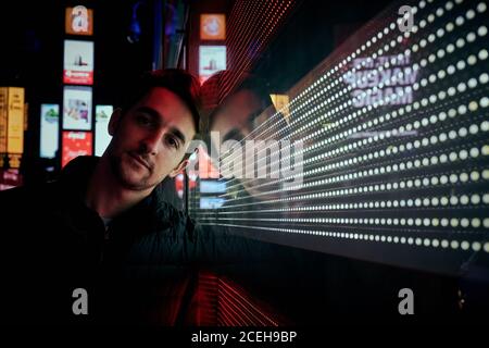 Young casual man leaning on glass shiny wall on street in night city looking at camera, New York Stock Photo