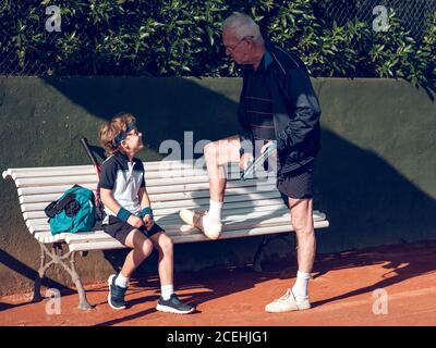 Aged grey male in sportswear with and little child with blue fillet and tennis rackets on seat talking in court in sunny day Stock Photo