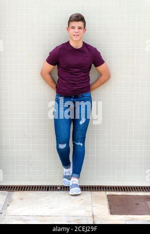 Handsome teenage man in casual outfit standing with hands behind back leaning on wall Stock Photo