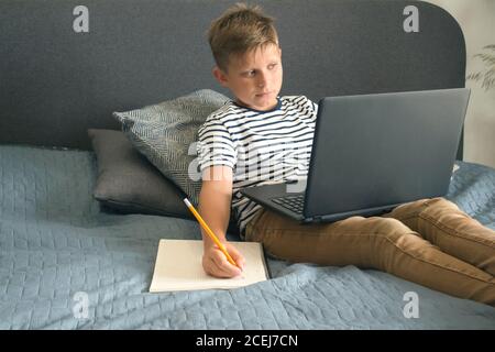 Front view portrait of diligent schoolboy writing or drawing, sitting on grey sofa while doing homework. Studying online during quarantine or personal Stock Photo