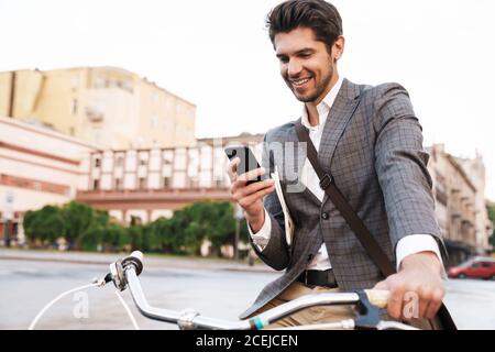 Image of happy handsome businessman in jacket smiling and using cellphone on bicycle at city street Stock Photo