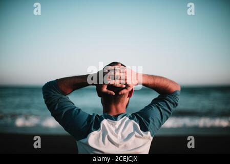 Back view of man standing with hands behind head and resting at the ocean. Stock Photo