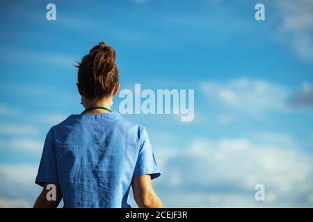 covid-19 pandemic. Seen from behind medical practitioner woman in scrubs outdoors in the city against sky. Stock Photo