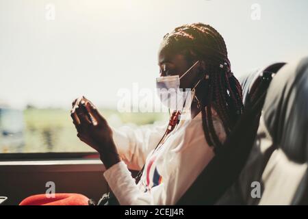 Side view of a young black woman tourist in a virus protective mask traveling in an intercity regular bus on the seat next to the window, she is watch Stock Photo