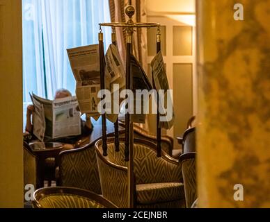 Seen from a different angle, the same Art Nouveau hall at Hotel Cresta Palace in Celerina, Switzerland, shows a guest reading behind the newspaper rack, surrounded by the original 1906 wood paneling. Stock Photo