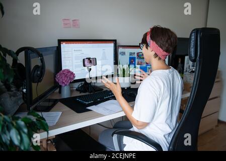 Back view creative blogger shooting video on phone camera sitting at her work desk with few monitors at home working space. E-learning. Remote work Stock Photo
