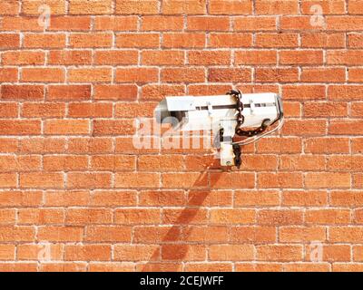 From below of white surveillance camera fixed with chain onto red brick wall in sunlight Stock Photo
