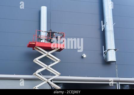 worker installs a ventilation system on the facade of a building using a scissor lift Stock Photo