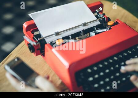 Crop close-up hands holding mobile phone and pen and using bright red typewriter Stock Photo